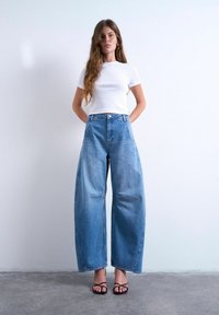 Femme debout contre un mur blanc, portant un haut blanc à manches courtes, un jean bleu à taille haute et jambes larges, et des sandales noires à lanières.