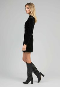 Femme portant une robe longue en velours noir à manches longues et des bottes à talons noires jusqu'aux genoux, se tenant de côté devant un fond gris clair uni.