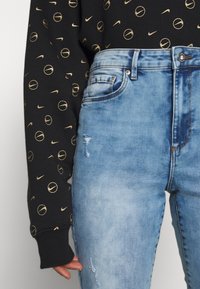 Sweat-shirt noir orné de cercles dorés et de logos Nike, associé à un jean bleu clair déchiré avec fermeture par bouton et poches.