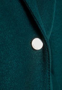 Blazer en laine teal avec une texture lisse, des revers aigus et un accent de bouton argenté brillant avec un détail embossé.