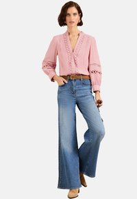 Blusa rosa con dettagli testurizzati e accenti in pizzo, abbinata a jeans svasati azzurri chiari e una cintura marrone, con calzature con tacco sobrie.