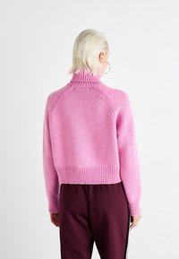 Rosa stickad tröja med hög krage och ribbat nederkant. Korta ärmar och texturerade mönster vid axelsömmarna. Kombinerad med mörka byxor.