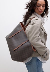 Sac fourre-tout à motif marron avec des anses en cuir marron. Texture lisse avec un design spacieux. Porté sur un manteau gris sur un mannequin.