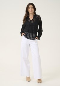 Black lace blouse with long sleeves and a scalloped hem, paired with wide-leg white pants and beige ankle boots.