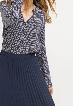 Frau trägt eine langärmelige, gemusterte, geknöpfte Bluse, die in einen hoch taillierten, marineblauen, plissierten Rock gesteckt ist, vor einem schlichten Hintergrund.