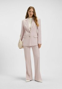 Hellrosa maßgeschneiderter Anzug mit strukturiertem Blazer und ausgestellten Hosen; verfügt über zwei Knöpfe, gekerbte Revers und seitliche Taschen. Begleitet von einer cremefarbenen Tasche.