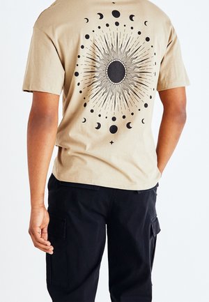 Person wearing beige t-shirt with black sun and moon phases graphic on back and black cargo pants, standing with one hand in pocket.