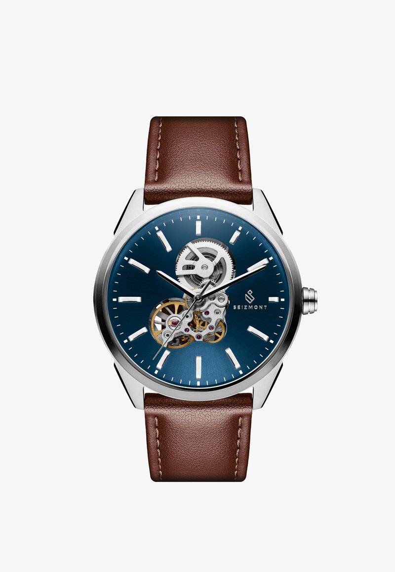 Mekanisk klokke med blå skive, transparent baklokk, sølvfarget rustfritt stålkasse og brun lærrem. Har minimale timemarkeringer.