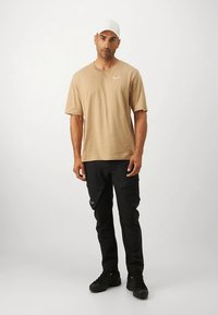 Bărbat purtând un tricou bej, pantaloni cargo negri, încălțăminte de drumeție negri și o șapcă albă, stând în fața unui fundal simplu, deschis la culoare.