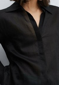 Camisa de lino negra con un collar clásico, botones visibles y una textura ligeramente translúcida. La tela tiene una caída suave y parece ligera.