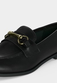 Mocassin en cuir noir avec détail mors en métal doré sur le dessus et semelle basse plate, vu en gros plan de profil.