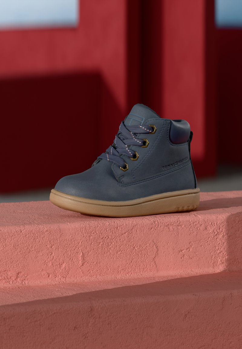 Botín de cuero azul hasta el tobillo con ojales de metal, suela de goma beige y cordones en contraste. Superficie texturada y cuello acolchado para mayor comodidad.