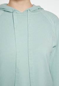 Felpa con cappuccio color turchese chiaro realizzata in tessuto a coste, con una patta frontale dettagliata da cuciture e maniche a raglan per un aspetto aderente.