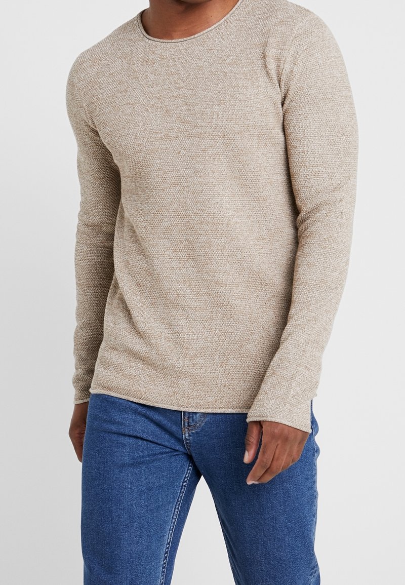 Man som bär en beige långärmad strukturerad tröja och blåa jeans, stående mot en enkel vit bakgrund.