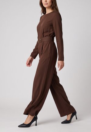 Frau läuft, trägt einen braunen langärmeligen Jumpsuit mit Gürtel und schwarze High Heels vor schlichtem grauem Hintergrund.