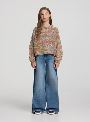 Giovane in piedi, indossando un maglione lavorato a maglia multicolore, jeans a gamba larga blu con pannelli laterali più scuri e sneakers marroni e bianche.