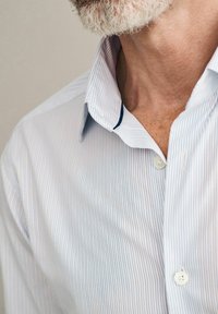 Svijetloplena i bijela prugasta košulja s razvedenim okovratnikom, kopčanjem na gumbi i suptilnim tamnoplavi akcentom duž ruba okovratnika.