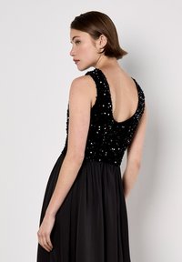 Abito nero con corpetto di paillettes e gonna fluida e testurizzata. Ha spalline larghe e un scollo sulla schiena poco profondo.