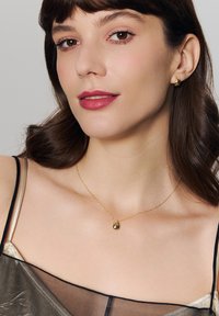 Złoty naszyjnik z wisiorkiem w kształcie kropli, noszony przez kobietę. Perłowe kolczyki dopełniają biżuterię. Gładka skóra na neutralnym tle.