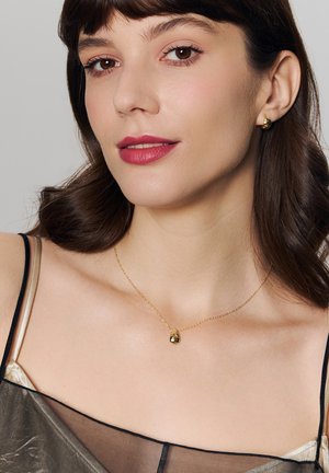 Złoty naszyjnik z wisiorkiem w kształcie kropli, noszony przez kobietę. Perłowe kolczyki dopełniają biżuterię. Gładka skóra na neutralnym tle.