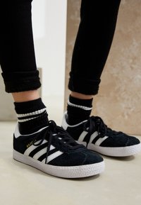 Sneakers in camoscio nero con dettagli a strisce bianche, suola testurizzata e lacci neri, abbinate a calzini neri e pantaloni neri risvoltati.