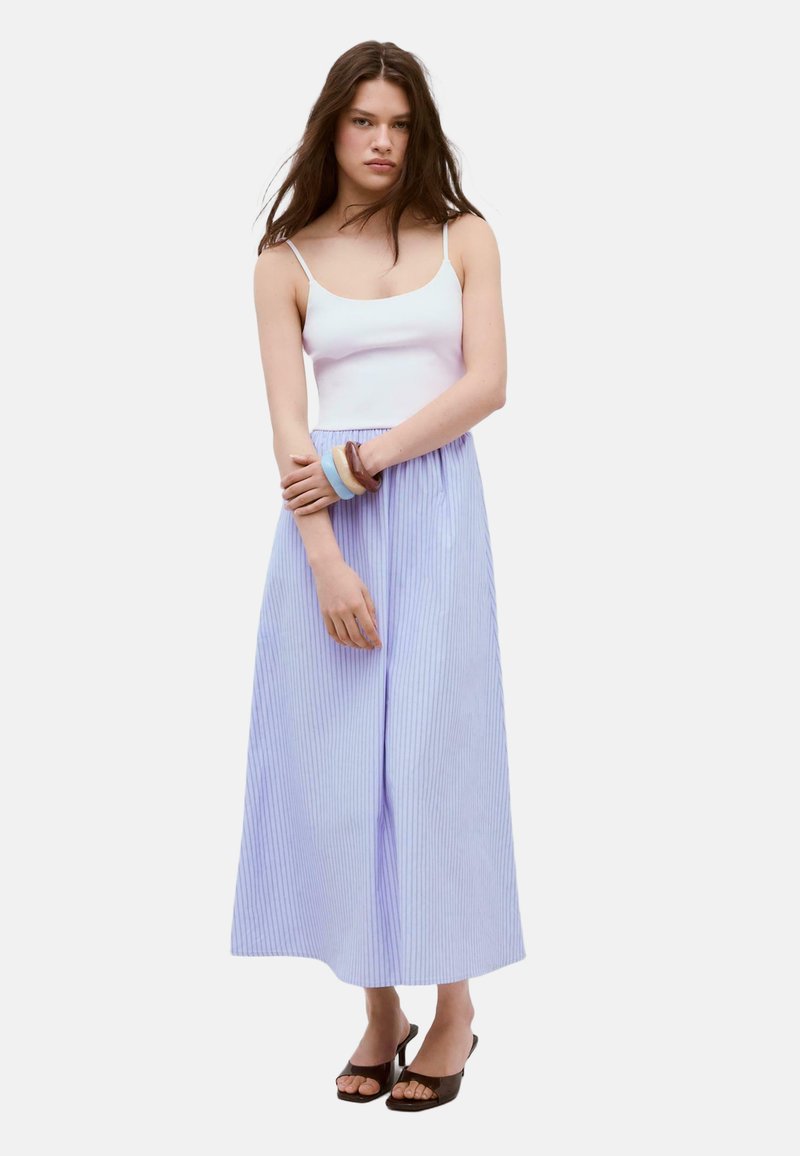 Jeune femme portant un débardeur blanc, un pantalon large plissé bleu clair, des sandales à talons noires et deux grands bracelets au poignet gauche.