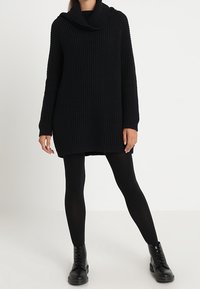 Schwarzer, gestrickter Oversize-Pullover mit überschnittenem Kragen, gerippten Bündchen und strukturiertem Muster, kombiniert mit schwarzen Leggings und Stiefeletten.