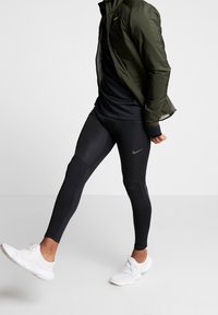 Mustat Nike-legginssit, joissa on istuva malli ja teksturoituja yksityiskohtia, yhdistettynä mustaan toppiin ja oliivinvihreään kevyttakkiin. Valkoiset lenkkarit.