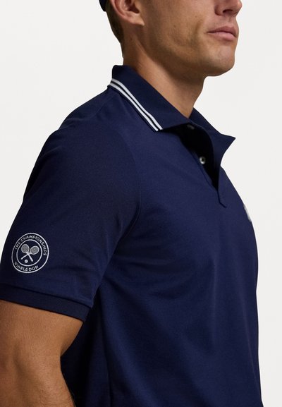 Polo shirt bleumarin cu guler striped alb, mâneci scurte și un logo circular Wimbledon pe mânecă stângă. Textură netedă, design ajustat.