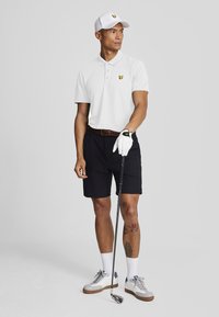 Uomo con berretto bianco, polo bianca, pantaloni corti neri e guanto da golf bianco, mentre tiene una mazza da golf, in piedi di fronte a uno sfondo grigio uniforme.