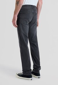 Mand iført sorte jeans med lige ben og sorte lave sneakers, står med ryggen til mod en ensfarvet grå baggrund.