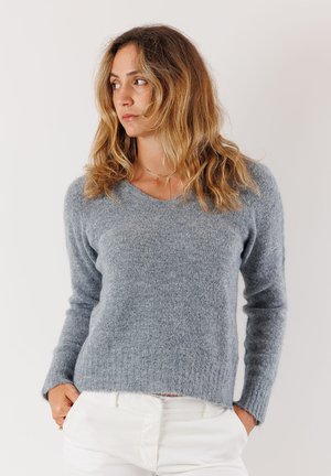Maglione grigio a maglia con scollatura a V e maniche lunghe, realizzato in materiale morbido. Presenta polsi a coste e un orlo rilassato. Indossato con pantaloni bianchi.