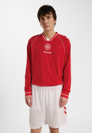 Junger Mann trägt ein rotes langärmeliges Sporttrikot mit weißem Logo und weißen Sportshorts vor einfarbigem Hintergrund.