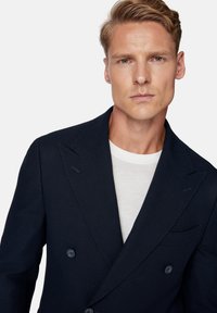 Granatowa, strukturalna marynarka dwurzędowa z wyłogami, dwoma przednimi kieszeniami i dopasowanymi guzikami, noszona na białej koszulce.