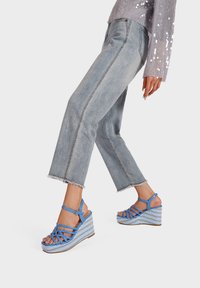 Sandali wedge blu con design a traverso e dettagli intrecciati sul tallone, abbinati a jeans chiari con orli sfilacciati.