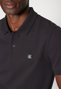 Homme portant un polo noir avec un petit patch logo "cK" sur la poitrine, sur fond uni clair.