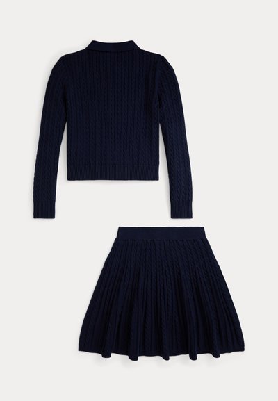 Σκούρο μπλε πλεκτό πουλόβερ με σχέδιο καλωδίων και γιακά, συνδυασμένο με φαρδιά φούστα που διαθέτει το ίδιο υφασμάτινο μοτίβο. Και τα δύο κομμάτια είναι ραμμένα με ραβδώσεις.