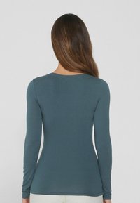 Langärmliges Oberteil in gedämpftem Teal, aus weichem, dehnbarem Stoff gefertigt. Verfügt über einen rundem Halsausschnitt und eine figurbetonte Passform, mit minimalen Nahtdetails.