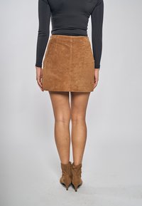 Ruskea mokkainen minihame, jossa on sileä pinta, yhdistettynä polven korkeisiin ruskeisiin korkosaappaisiin, nähtynä takaa neutraalia taustaa vasten.