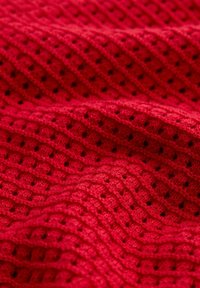 Tissu tricoté rouge avec un motif texturé de crêtes diagonales et de petits trous, montré en gros plan avec des détails visibles des fibres.
