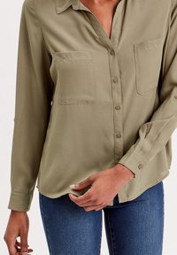 Chemise boutonnée vert olive avec col, deux poches avant et manches longues avec poignets boutonnés, présentant un ourlet légèrement incurvé.