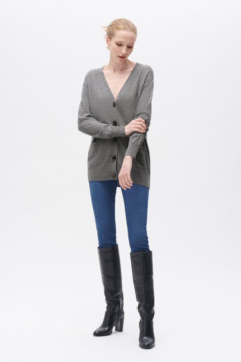 Grauer Cardigan mit V-Ausschnitt, ausgestattet mit großen braunen Knöpfen und einer lockeren Passform. Kombiniert mit blauen Jeans und schwarzen kniehohen Stiefeln.