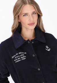 Marine korduroy jack met een kraag, voorzien van zilveren drukknoopjes en een geborduurde ankerdetail, samen met tekst aan de linkerkant.