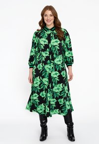 Knopfkleid mit schwarzem Grund und grünem Blumenmuster. Mit Kragen und 3/4-Ärmeln, kombiniert mit kniehohen schwarzen Stiefeln.
