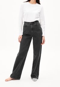 Zwart denim jeans met hoge taille en rechte pijpen, voorzien van zijzakken en riemlussen; gecombineerd met een wit shirt met lange mouwen.