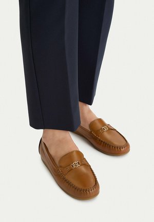 Pies con mocasines de cuero marrón con detalle de hebilla dorada, combinados con pantalones azul marino recortados sobre un fondo claro.