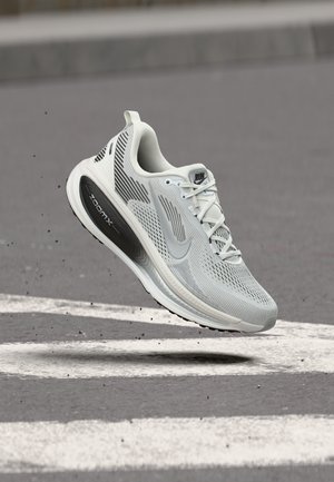 Hellgrauer Sportschuh mit schwarzer "ZoomX"-Sohle, weißen Schnürsenkeln und "RUN"-Aufschrift, schwebt in der Luft über asphaltiertem Untergrund mit weißen Streifen.