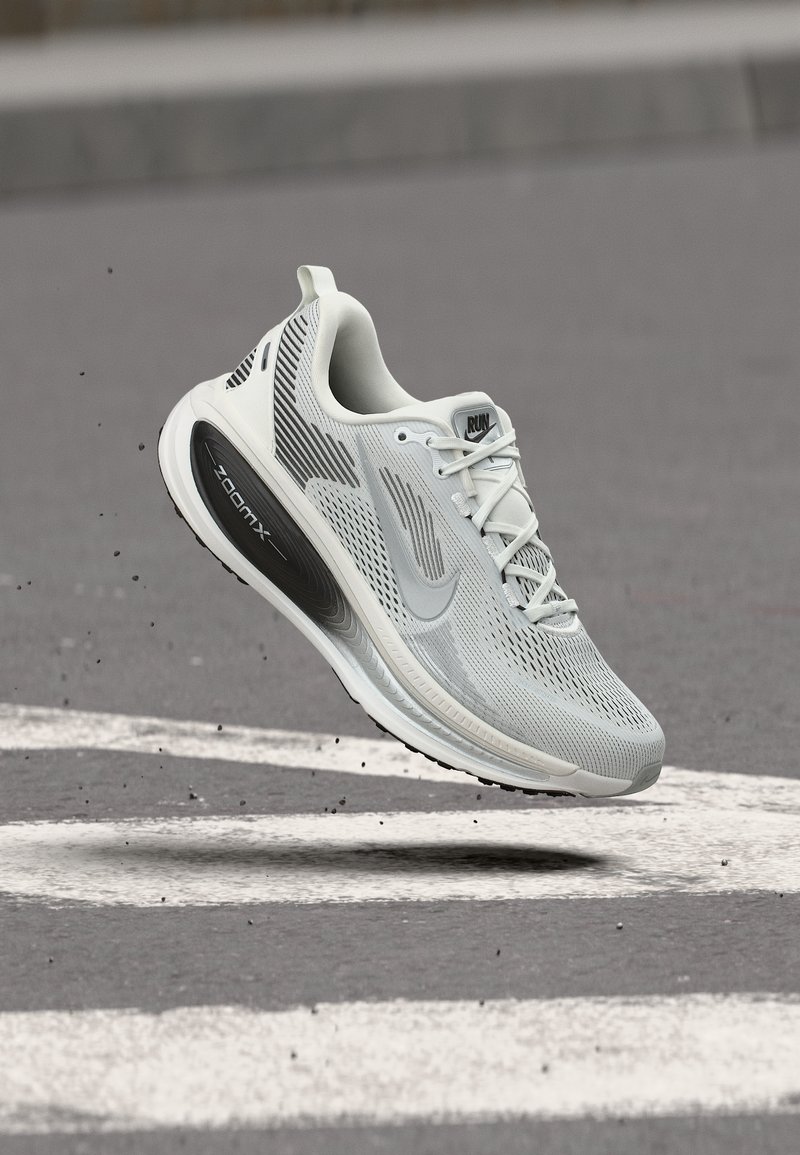 Αθλητικό παπούτσι ανοιχτό γκρι με μαύρη σόλα "ZoomX", λευκά κορδόνια και ετικέτα "RUN", εμφανιζόμενο στον αέρα πάνω από άσφαλτο με λευκές ρίγες.