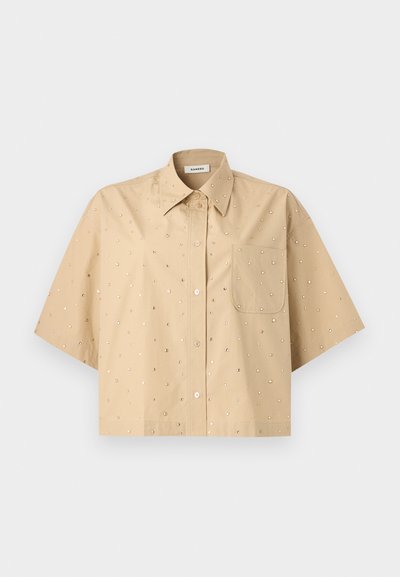 Μπεζ πουκάμισο με κοντά μανίκια και κουμπιά, μυτερό γιακά, τσέπη στο στήθος και μικρές ανακλαστικές κουκκίδες διάσπαρτες στο ύφασμα.