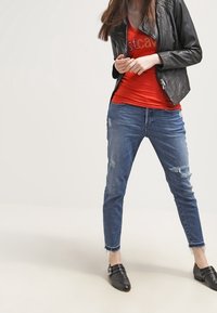 Giubbotto in pelle nera da motociclista, top rosso aderente con strass, jeans blu scoloriti con orli sfilacciati e mocassini neri.